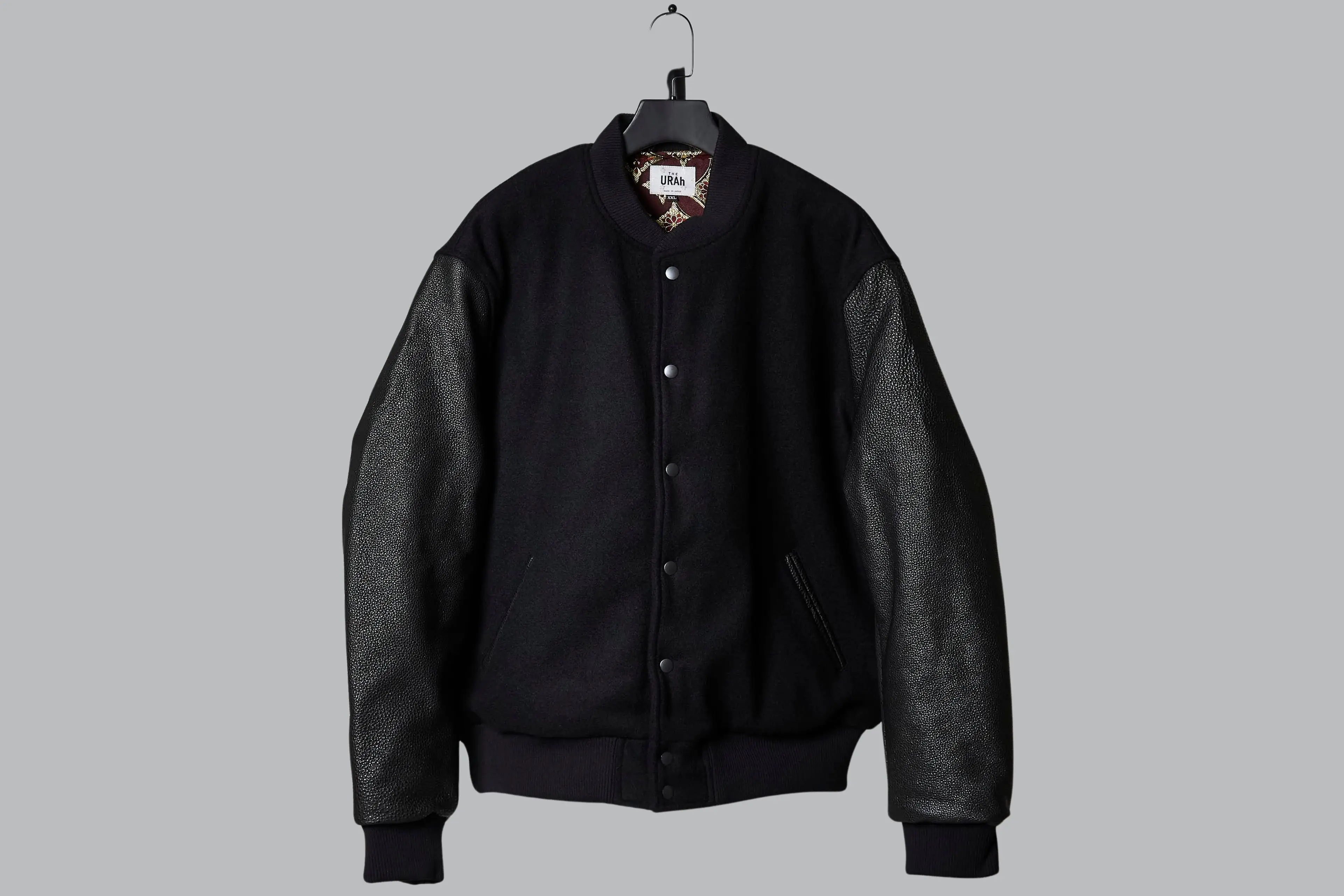 A black varsity jacket on a hanger against a grey ground: Bishu melton wool body, urushi-lacquered kurozan leather sleeves, black ribbing, and a glimpse of the crimson and gold lining at the collar.