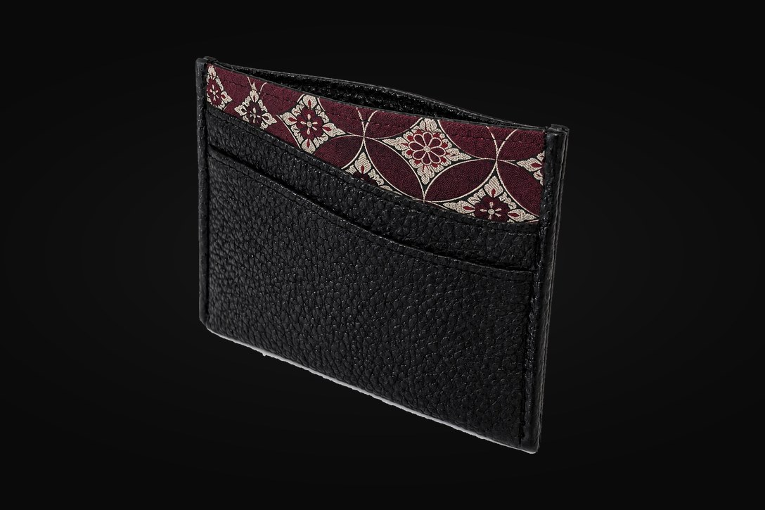 A kurozan leather card case standing at an angle, its card slot open on a crimson and gold shippō-patterned lining, against black.